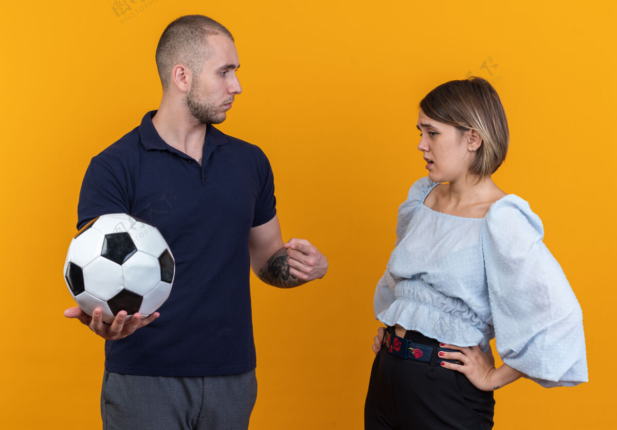 足球年轻漂亮的情侣穿着休闲服拿着足球的男人看着他困惑不安的女友站着漂亮随意站立