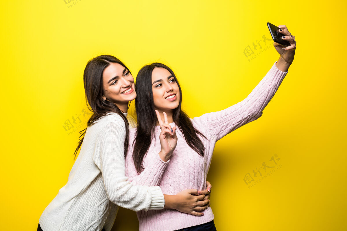 时尚两个穿着毛衣的可爱女孩站在黄色墙壁上自拍的照片微笑朋友照片