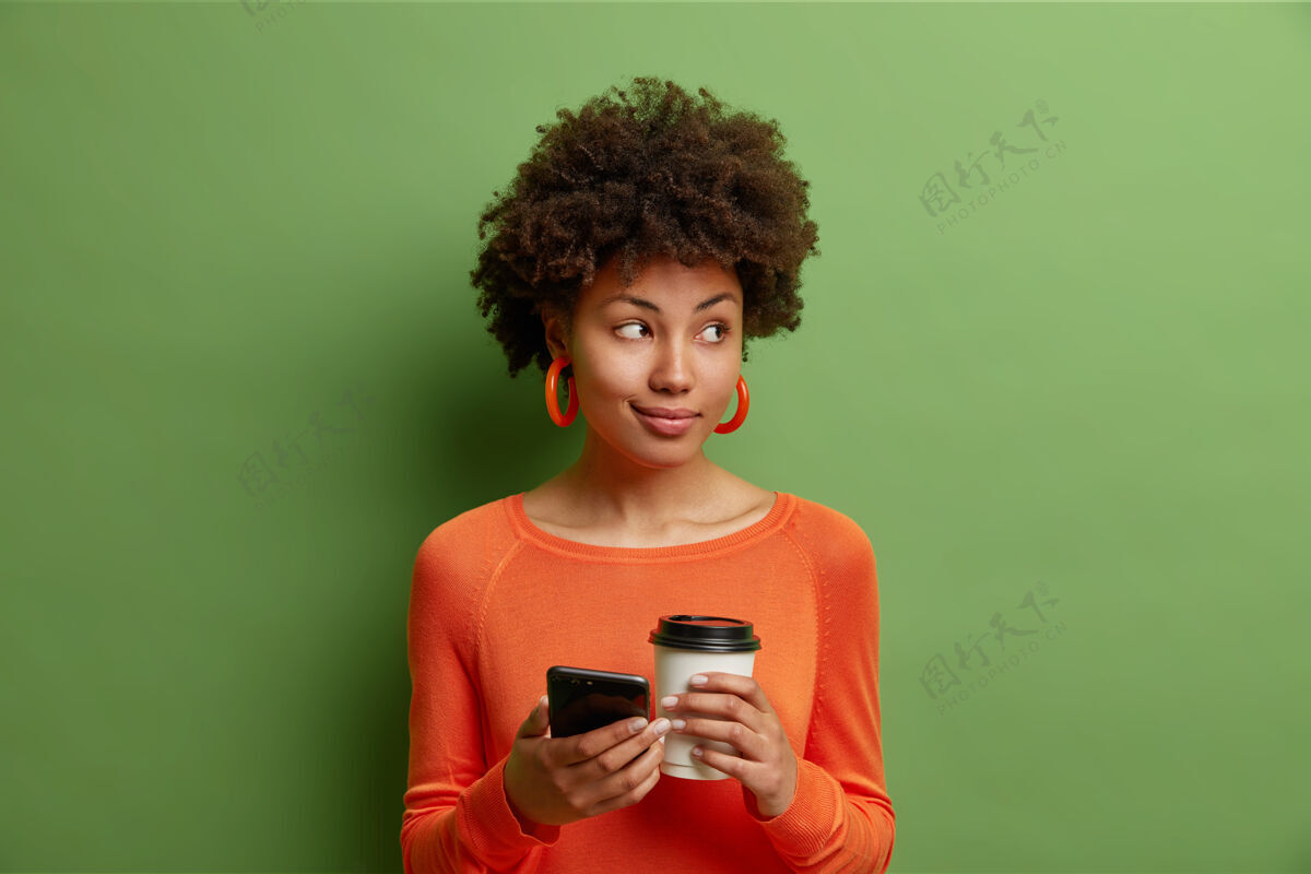 室内卷发美女捧着一杯美味的咖啡 智能手机穿着休闲的橙色毛衣隔着鲜艳的绿色墙壁传递信息咖啡无线科技