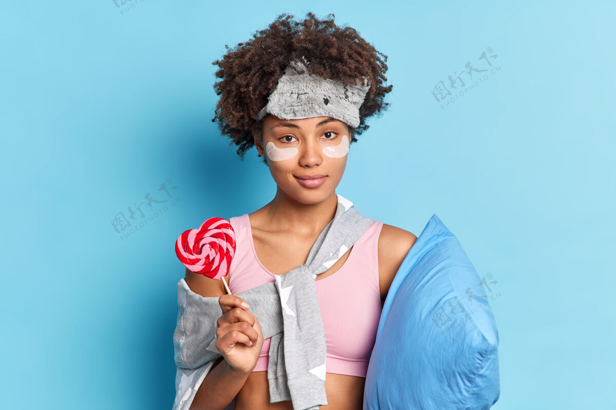 感觉严肃自信的美丽女人的画像拿着甜甜的糖果在健康的睡眠后醒来穿着黑色的衣服 枕头靠在蓝色的墙壁上枕头睡衣早晨