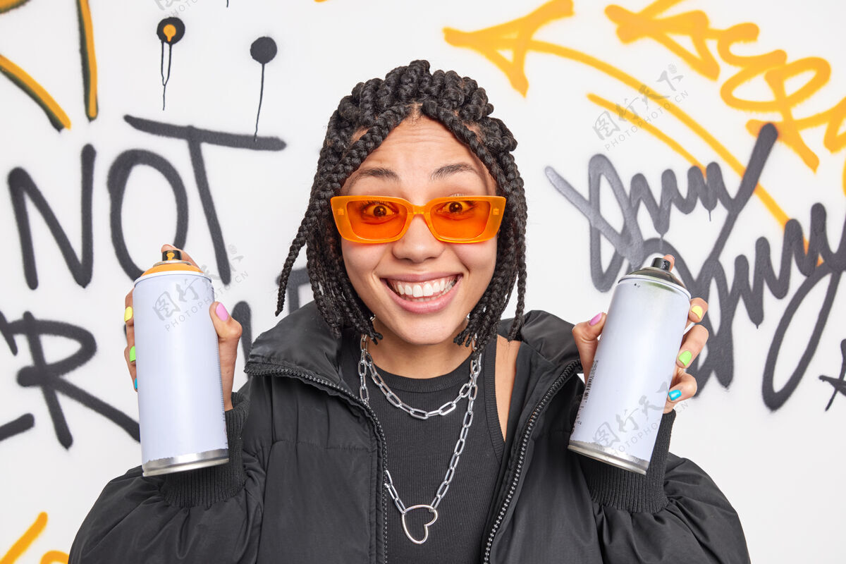 涂鸦快乐的千禧一代女人带着辫子拿着两个喷雾瓶正在创作街头艺术家涂鸦穿着时髦的衣服橙色太阳镜属于帮派青年高兴青少年