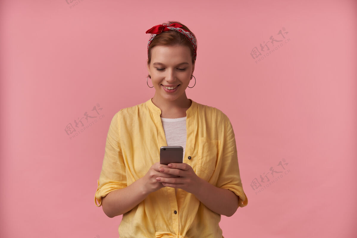 欢呼微笑着穿着黄色衬衫 头上戴着头巾的年轻女子站在粉红色的墙上用手机人物手机女士