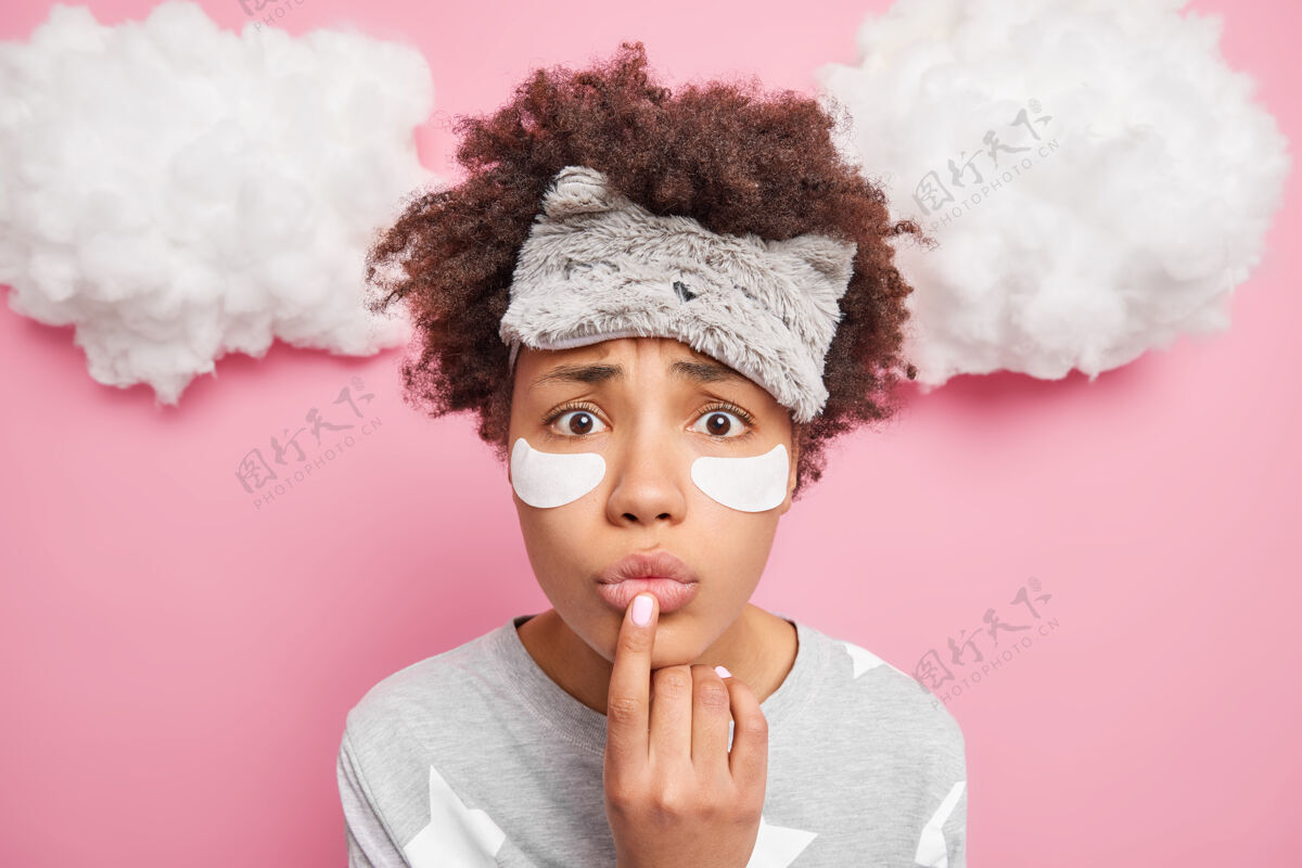 穿着紧张的非裔美国妇女的特写镜头 手指放在折叠的嘴唇上 穿着睡衣 额头戴着睡眠面罩 隔着粉色的墙壁 上面是白云女性室内醒着