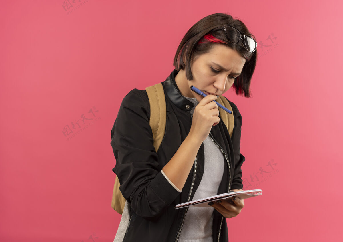 脸体贴的年轻女学生头戴眼镜 背着书包 手里拿着笔和笔记本 看着笔记本 用手指抚摸着嘴唇 把手指孤立在粉红色的墙上触摸女孩拿着