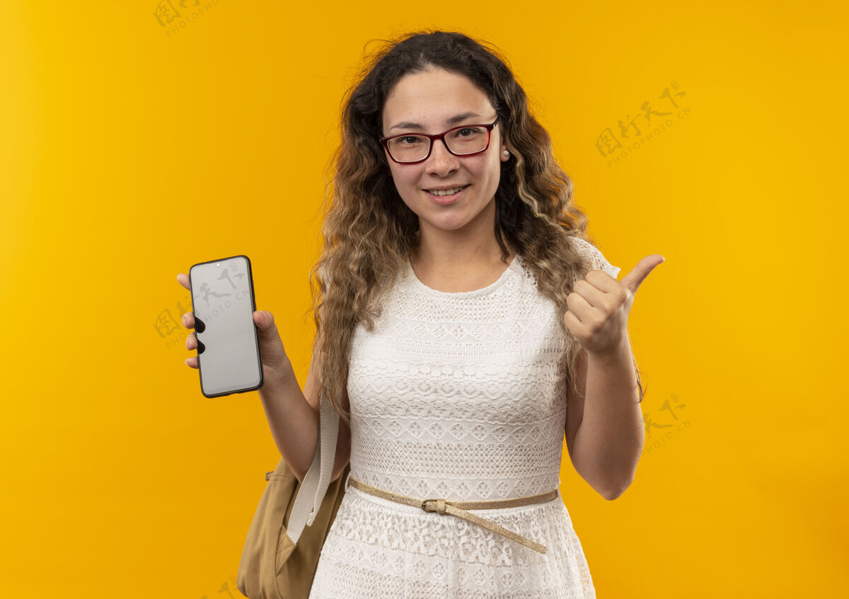 眼镜微笑着的年轻漂亮的女学生戴着眼镜 背着书包 拿着手机 大拇指向上竖起 孤零零地站在黄色的墙上电话背部人