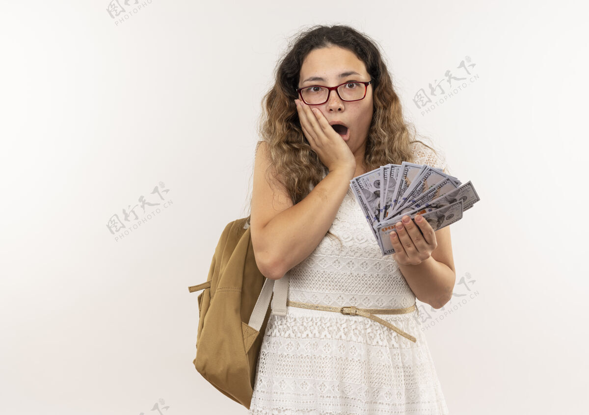脸惊讶的年轻漂亮的女学生戴着眼镜背着包拿着钱手放在脸上隔离在白墙上市民人穿