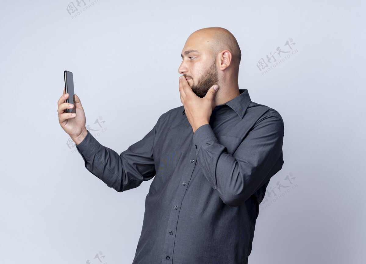 人体贴的年轻秃头呼叫中心的男人手放在嘴上拿着手机看着隔离在白墙上人姿势秃头