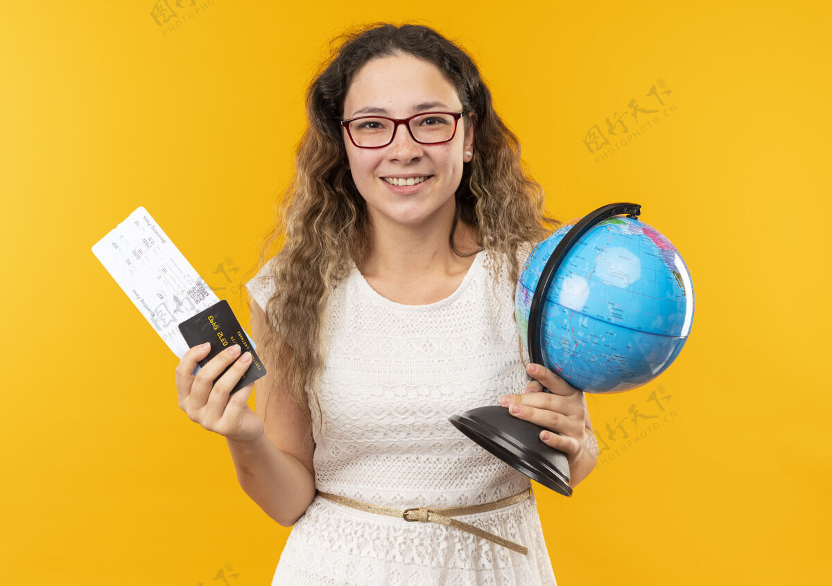 地球微笑的年轻漂亮的女学生戴着眼镜拿着机票 信用卡和地球仪孤立在黄色的墙上票微笑信用