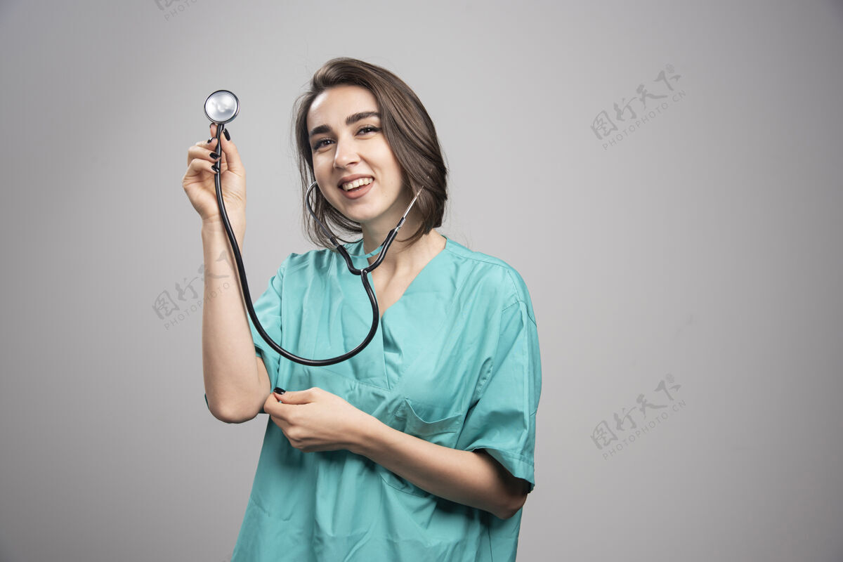 年轻快乐医生在灰色背景上展示听诊器高质量照片医生制服工作