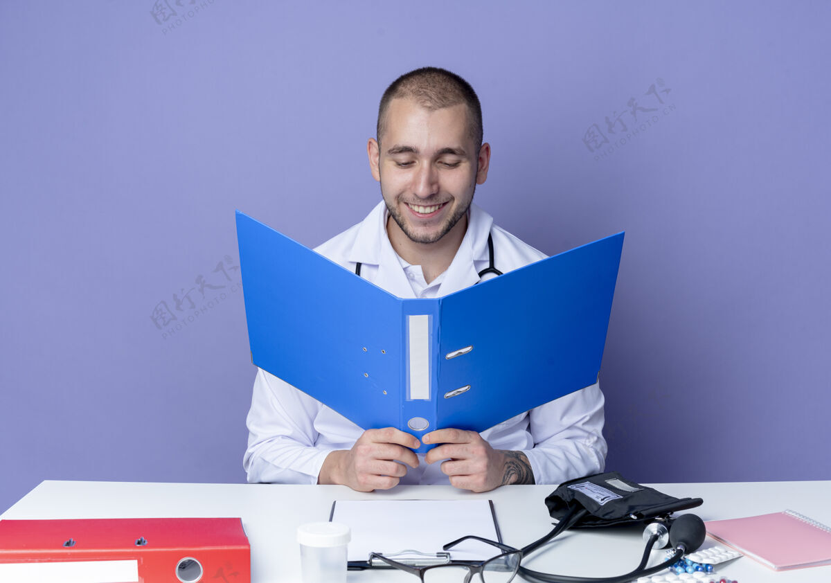 脸面带微笑的年轻男医生穿着医用长袍和听诊器坐在办公桌旁 手里拿着工作工具 看着隔离在紫色墙上的文件夹桌子文件夹表情