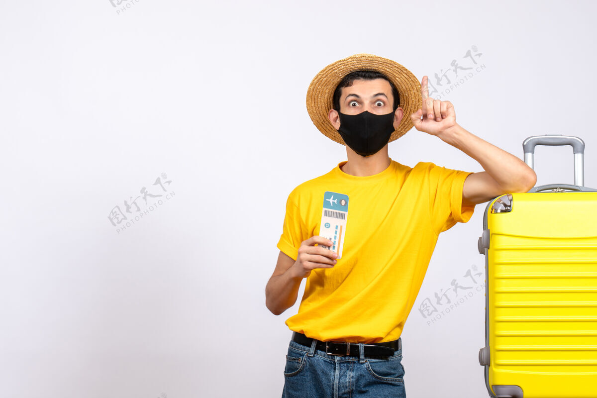 睁大眼睛正面图：戴草帽的大眼睛年轻人站在黄色手提箱旁 拿着旅行票 手指向上视图人吸管