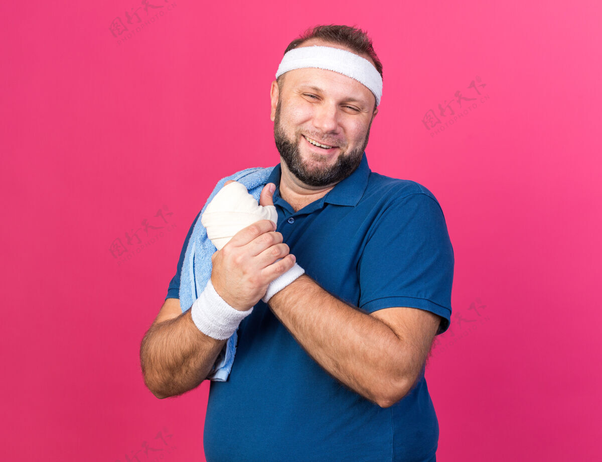 男人面带微笑的成年斯拉夫运动型男子 肩上披着毛巾 头上戴着头带 手腕上戴着手环 手被隔离在粉色的墙上 留着复制空间斯拉夫肩膀腕带