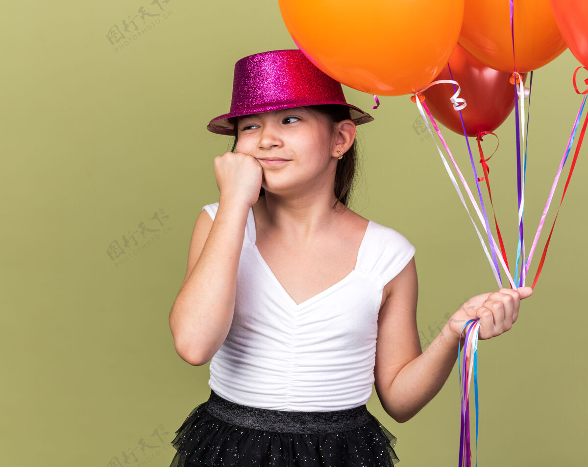 拳头高兴的年轻白人女孩 戴着紫色派对帽 手里拿着氦气球 把拳头放在脸上 看着橄榄绿墙壁上孤立的一面 还有复制空间气球白种人橄榄