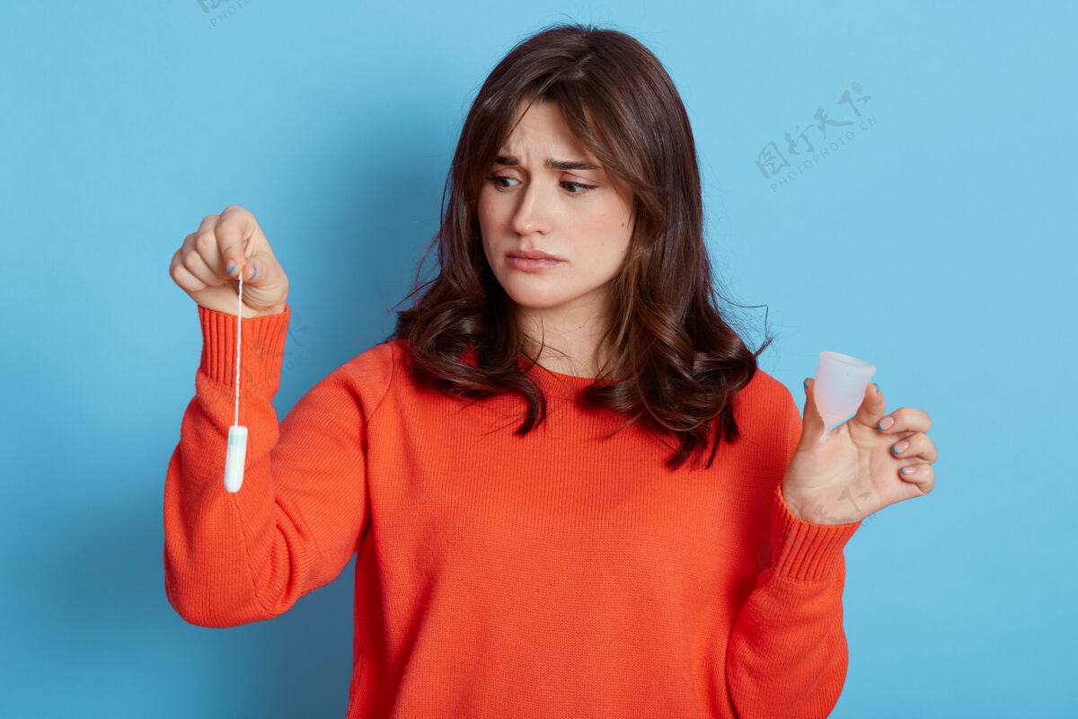 烦恼愁眉苦脸的黑发女穿着橘色毛衣手里拿着女人的卫生用品方法卫生棉条保护