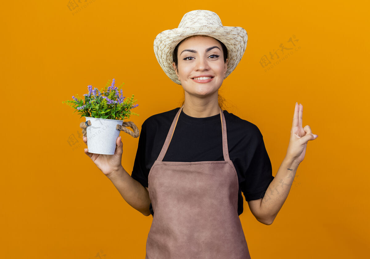 信心年轻漂亮的女园丁 围着围裙 戴着帽子 手里拿着盆栽植物 微笑着自信地看着前面站在橙色墙上的二号人物站着花园微笑
