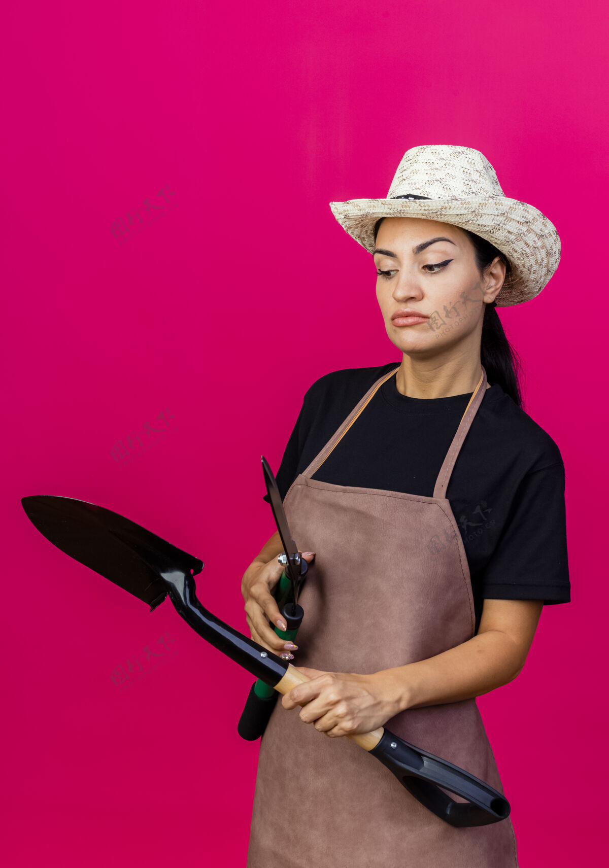 年轻年轻漂亮的女园丁围着围裙 戴着帽子 手里拿着树篱剪和铲子 严肃的脸站在粉红色的墙上望着一边花园剪子表情