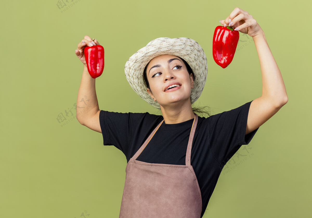 光年轻漂亮的女园丁 围着围裙 戴着帽子 手里拿着红甜椒 站在浅绿的墙上 欢快地笑着抱着美丽欢呼