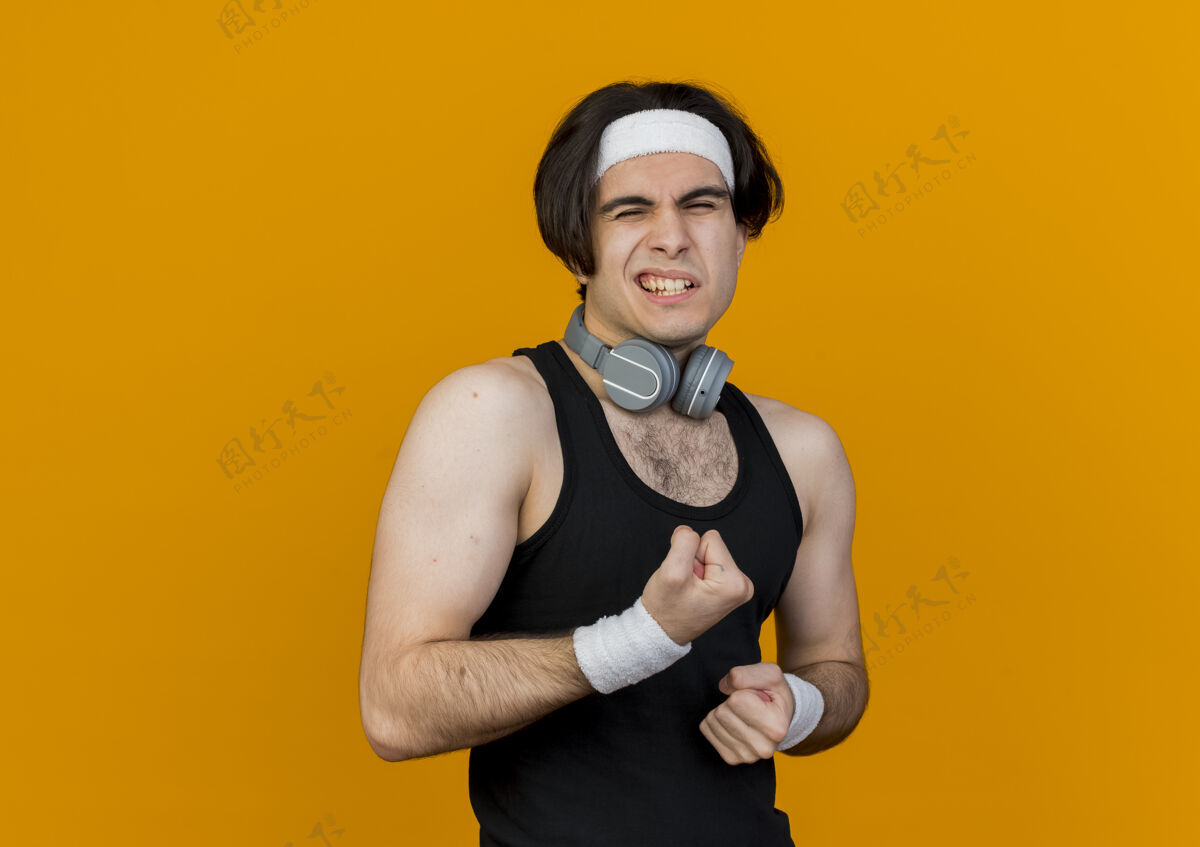 头带穿着运动服 头戴耳机 脖子上戴着头带的年轻人站在橘色的墙上看着紧握的拳头姿势人拳头