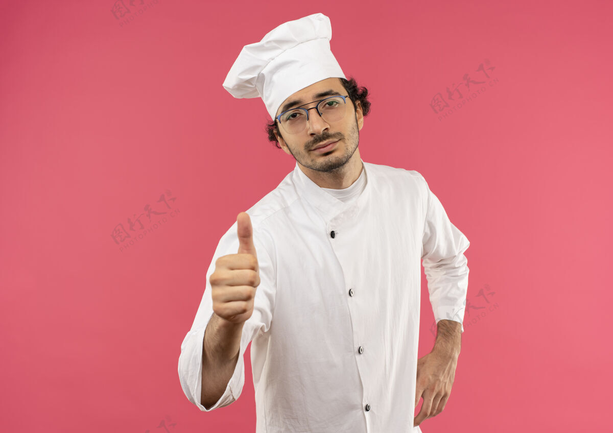 手自信的年轻男厨师穿着厨师制服 戴着眼镜 手放在臀部 拇指朝上烹饪背景自信