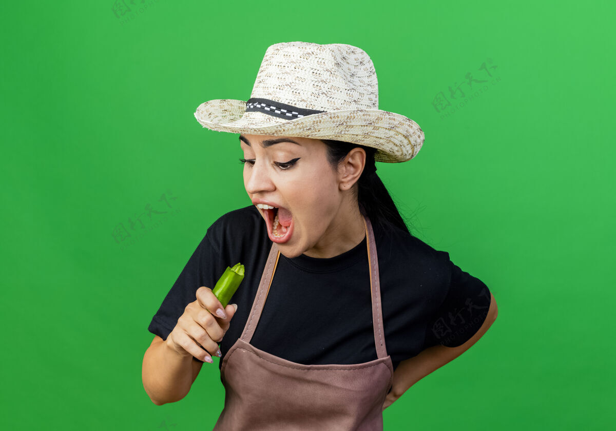 围裙年轻漂亮的女园丁站在绿色的墙上 围着围裙 戴着帽子 手里拿着绿色的辣椒试图咬它咬市民脸