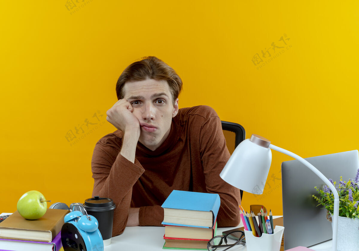 男孩看着摄像机年轻的学生男孩正坐在书桌旁 头戴在手腕上坐着黄色背景