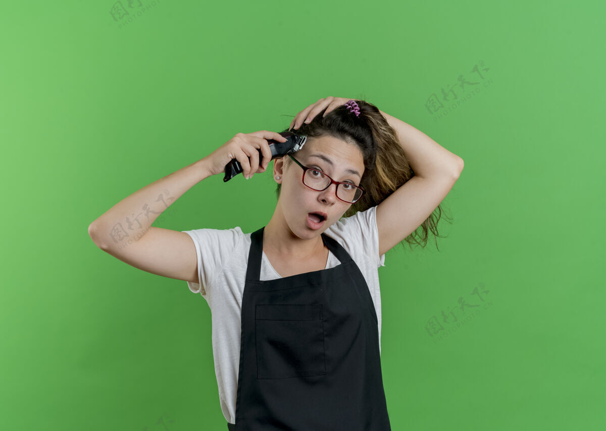 人年轻的专业美发师穿着围裙剪着头发的女人站在绿色的墙前惊讶地看着前面理发师女人表情