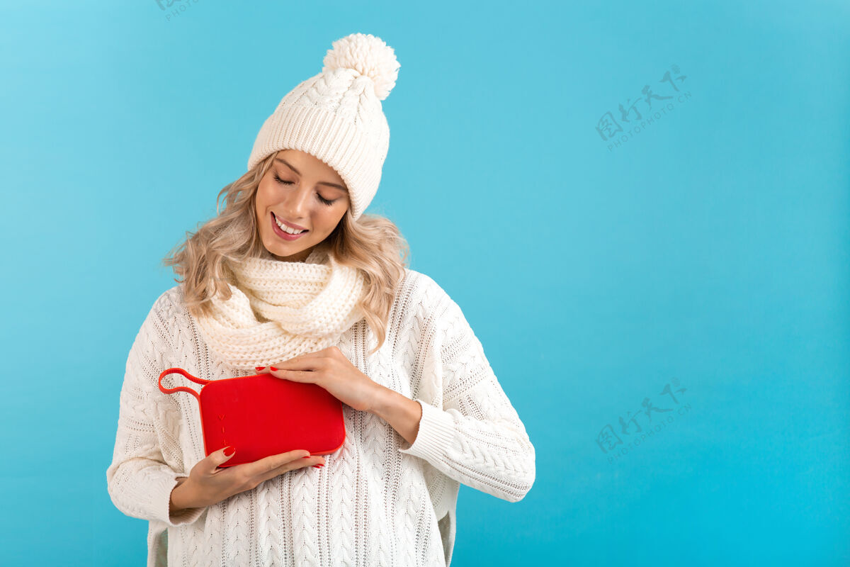 女士时髦的金发微笑的漂亮年轻女子手持无线扬声器听音乐快乐地穿着白色毛衣和针织帽子摆在蓝色的姿势乐趣模型无线
