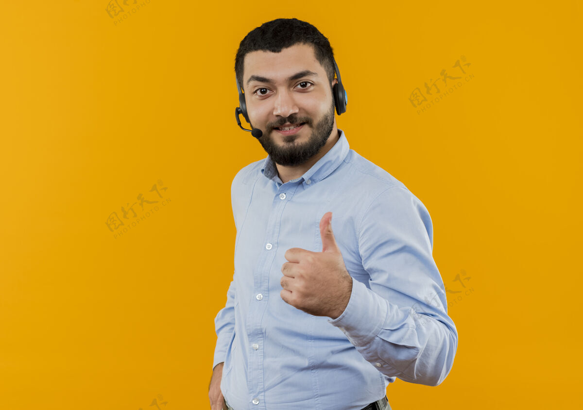 人一个留着胡子的年轻人 穿着蓝色衬衫 戴着耳机 拿着麦克风 面带微笑地站在橙色的墙上竖起大拇指微笑麦克风胡须