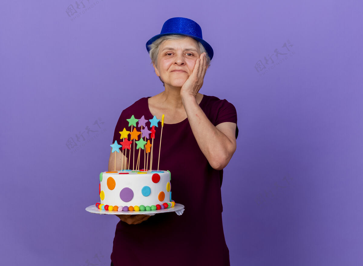 蛋糕戴派对帽的老太太高兴地把手放在脸上 把生日蛋糕孤立地放在紫色的墙上市民人女人