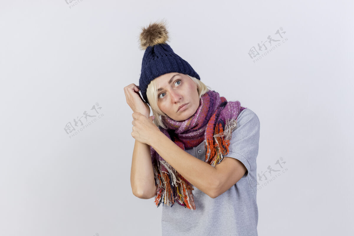 围巾自信的年轻金发病女人戴着冬天的帽子和围巾抱着胳膊看着隔离在白墙上的一面脸姿势举行