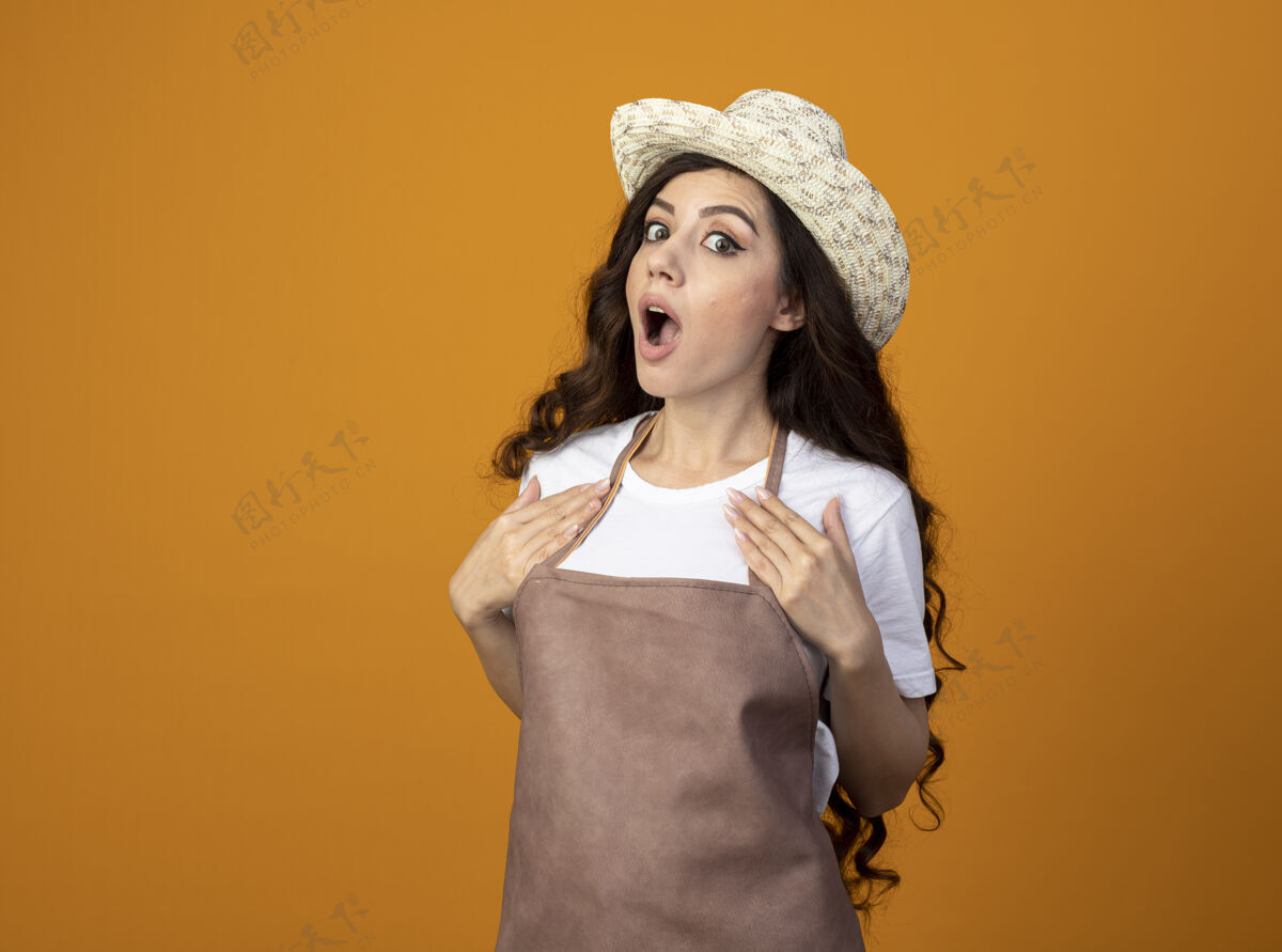 惊喜穿着制服的年轻女园丁惊讶地戴着园艺帽把手放在隔离在橙色墙上的胸口上制服穿着年轻人
