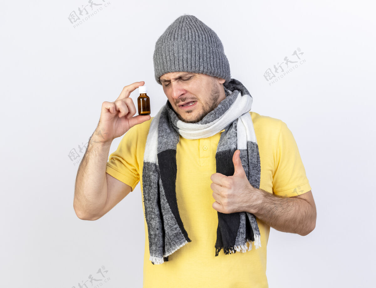 表情戴着冬天帽子和围巾的年轻金发病患拿着玻璃瓶里的药 竖起大拇指孤零零地站在白墙上人帽子穿着