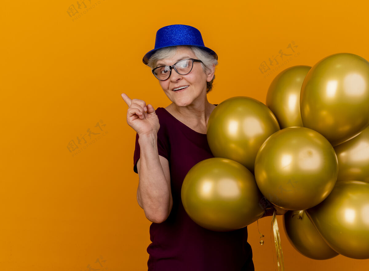 指向戴着聚会帽 戴着眼镜的老太太高兴地站在那里 氦气球指着橙色墙上孤立的一面女人脸请