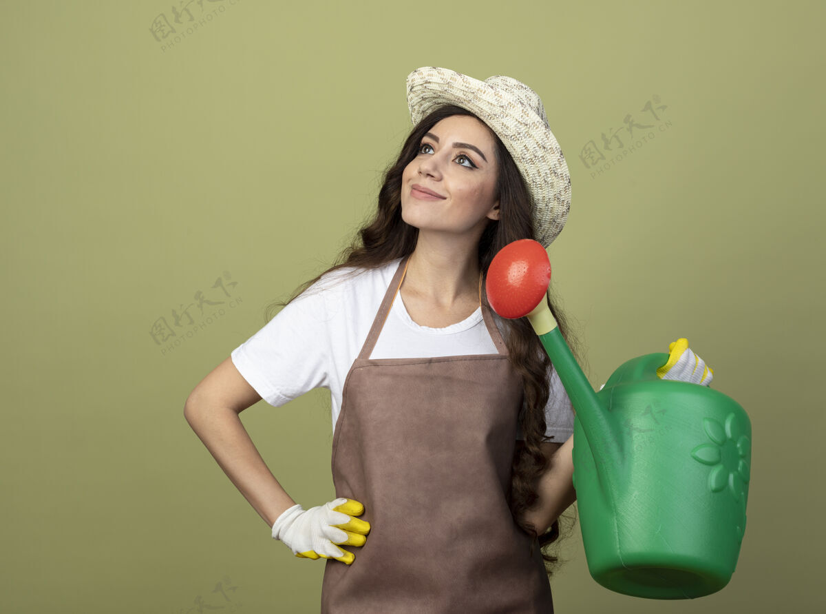 帽子身着制服 戴着园艺帽和手套的年轻女园丁高兴地拿着浇水罐 看着橄榄绿墙上孤立的一面手套穿着女性