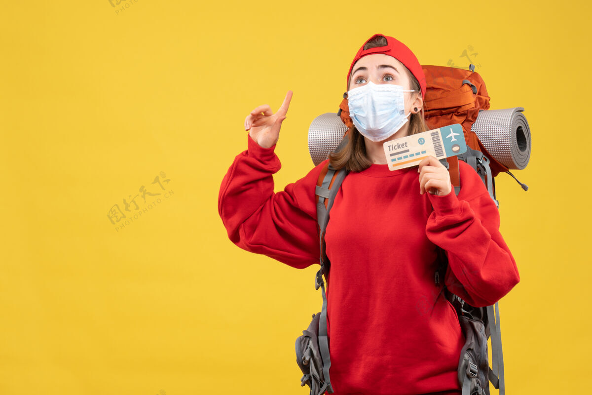 战争正面图：年轻的女游客背着背包 戴着口罩 手举旅行票 手指向上票手指背包