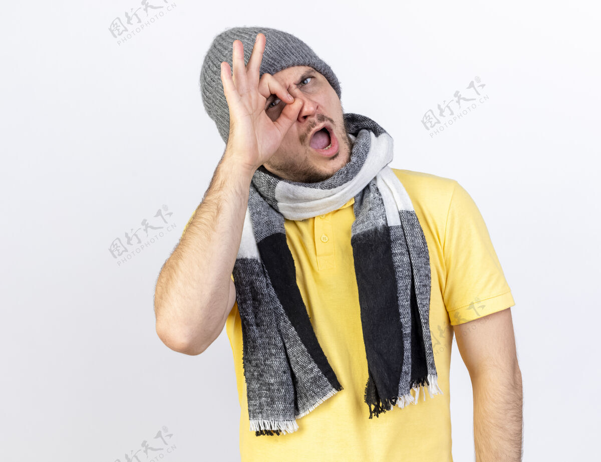围巾愤怒的年轻金发病男子戴着冬天的帽子和围巾看着前面通过手指隔离在白墙上手指愤怒表情