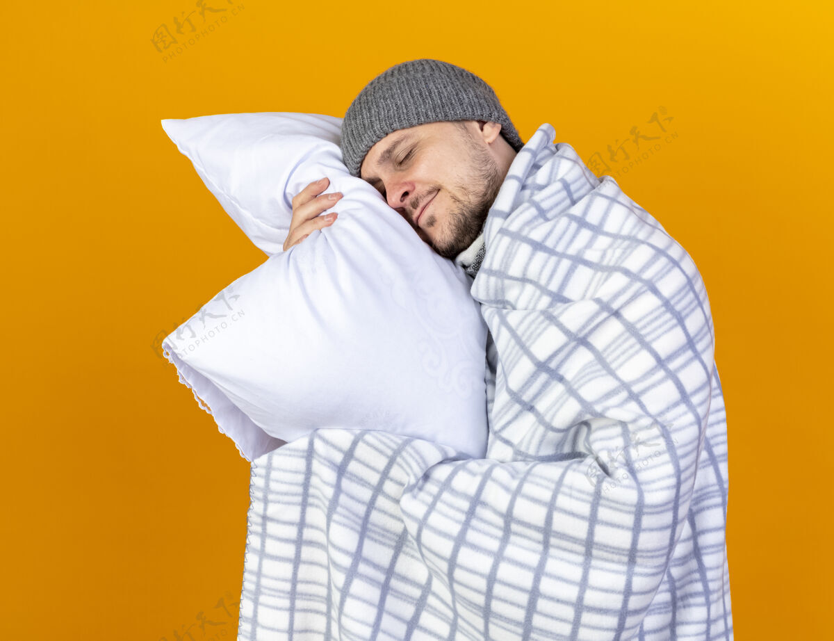 脸高兴的年轻金发病男子戴着冬天的帽子和围巾包裹在格子布举行和把头放在枕头上隔离在橙色的墙壁人金发年轻人