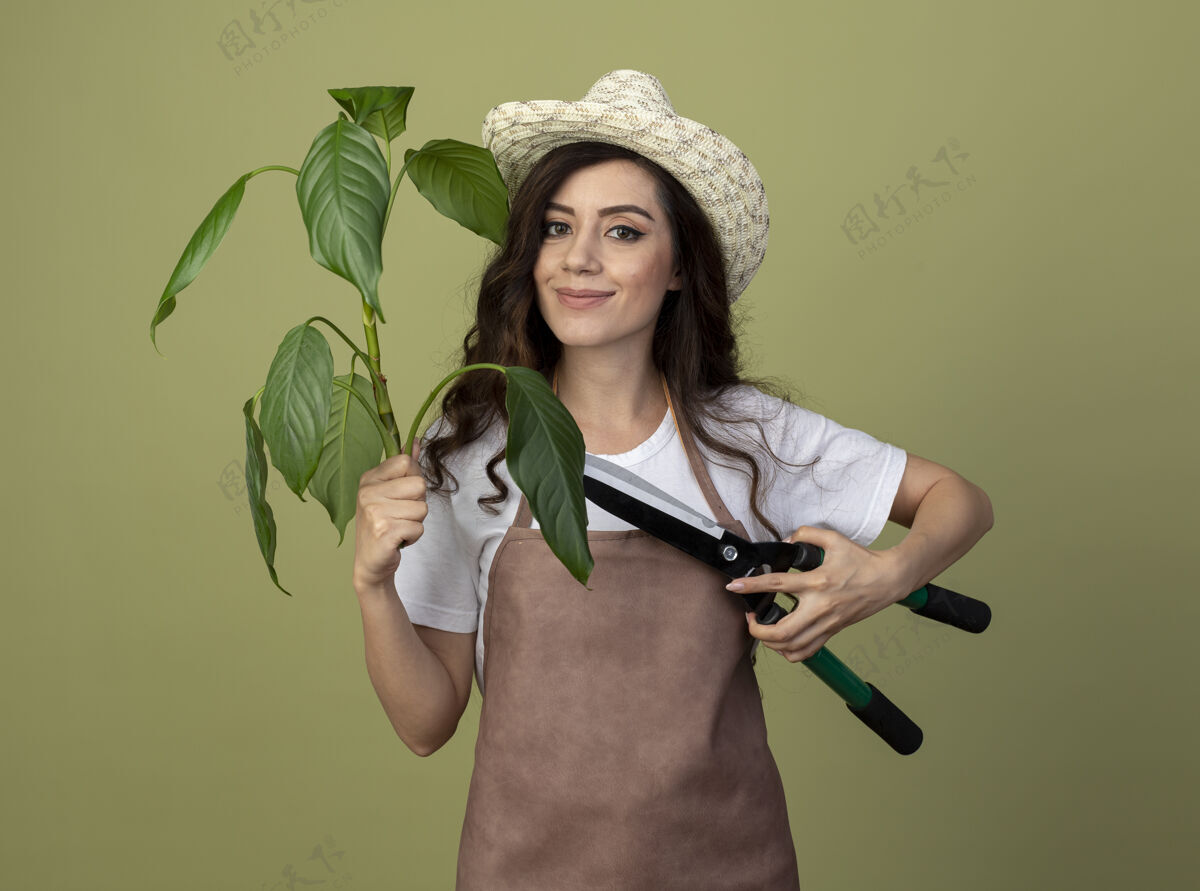脸身着制服 戴着园艺帽的年轻女园丁高兴地把植物和园艺剪隔离在橄榄绿的墙上园艺市民帽子