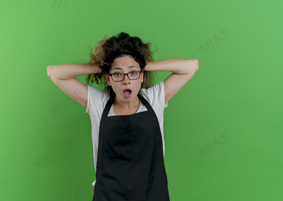 公民年轻的专业美发师围着围裙看着前面的女人惊慌失措地拉着头发站在绿色的墙上拉姿势围裙