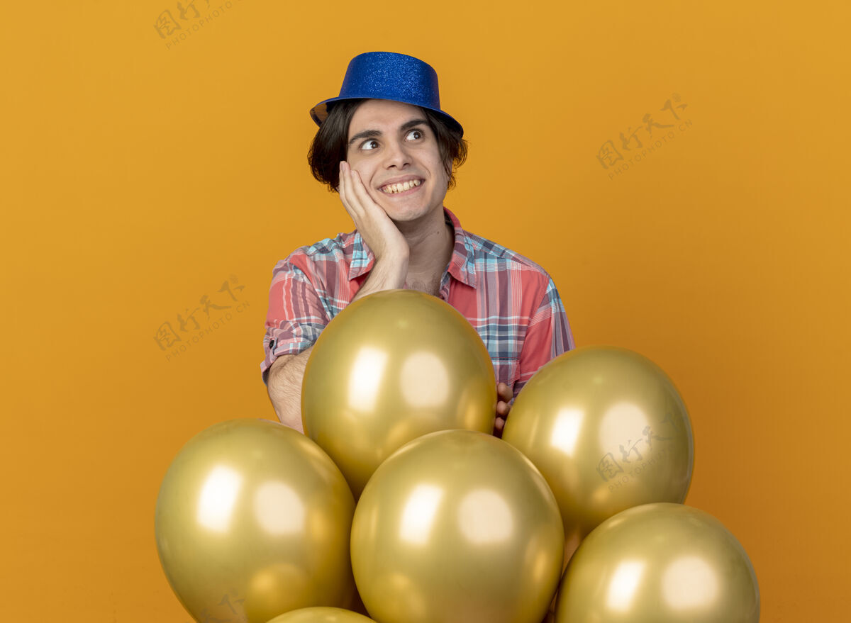 站戴着蓝色派对帽的帅哥兴奋地把手放在脸上 站在那里 手里拿着氦气球 看着橙色墙上孤立的一面脸侧线人