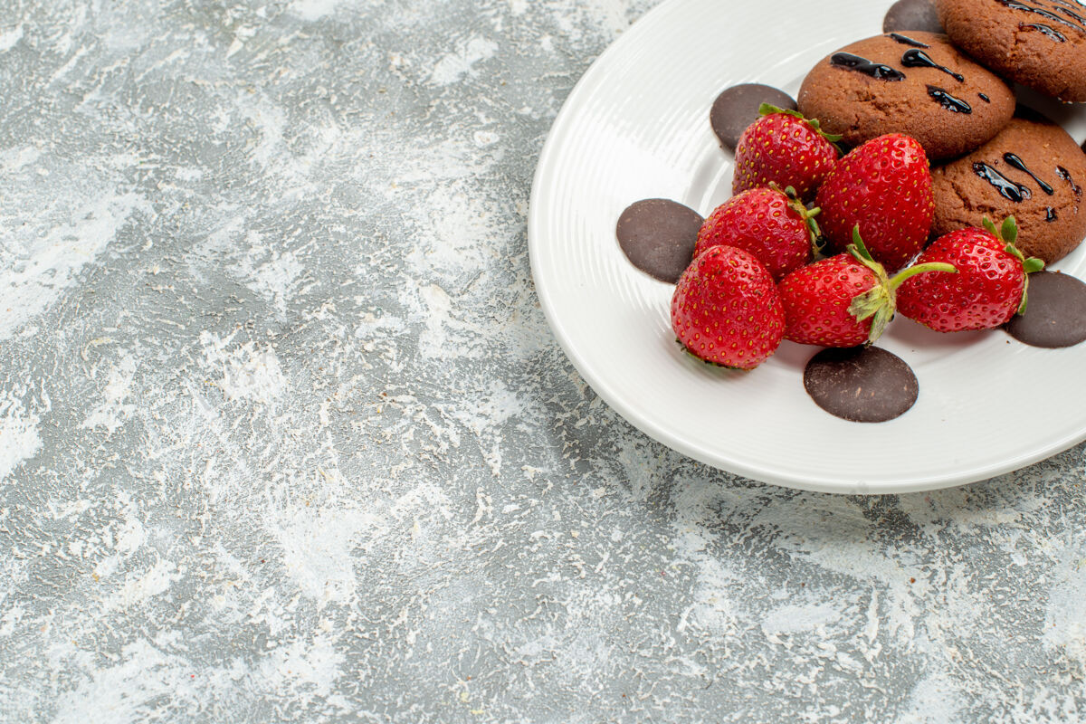 草莓上半部分查看巧克力饼干草莓和圆形巧克力在白色椭圆形盘子在顶部的灰白色地面右上角磨碎的可食用水果水果