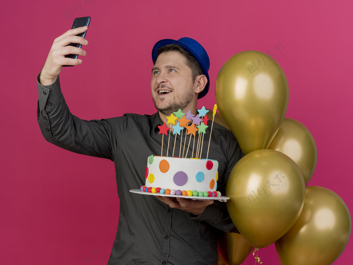 拿着微笑着戴着蓝色帽子的年轻人站在气球旁拿着蛋糕 在粉红色的天空中自拍帽子派对抱着
