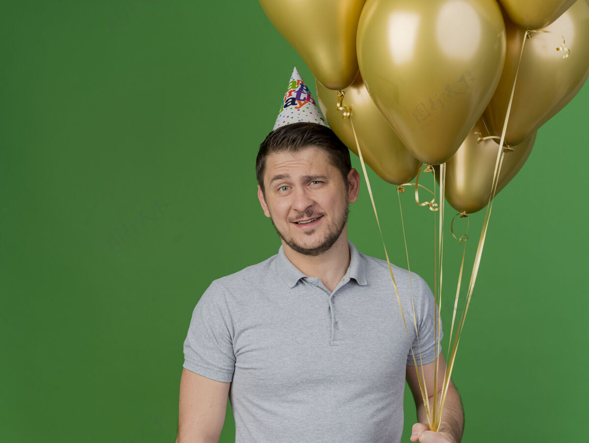 戴着高兴的年轻人穿着灰色衬衫和生日帽举行聚会举行气球隔离在绿色气球生日小伙子