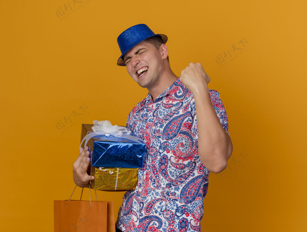 年轻快乐的年轻人闭着眼睛戴着蓝色的帽子 拿着礼品盒和袋子 在橙色上显示“是”的手势手势派对礼物
