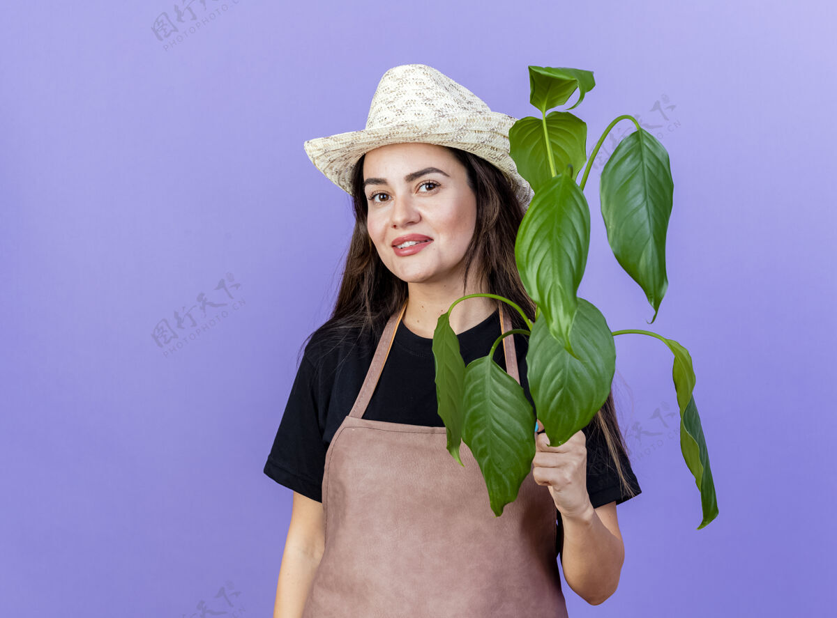 制服微笑着美丽的园丁女孩穿着制服戴着园艺帽拿着隔离在蓝色的植物帽子园艺穿