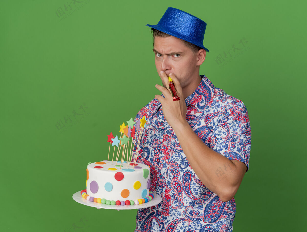 穿一个戴着蓝帽子 拿着蛋糕 吹着绿色的吹风机的严格的年轻人帽子吹绿色