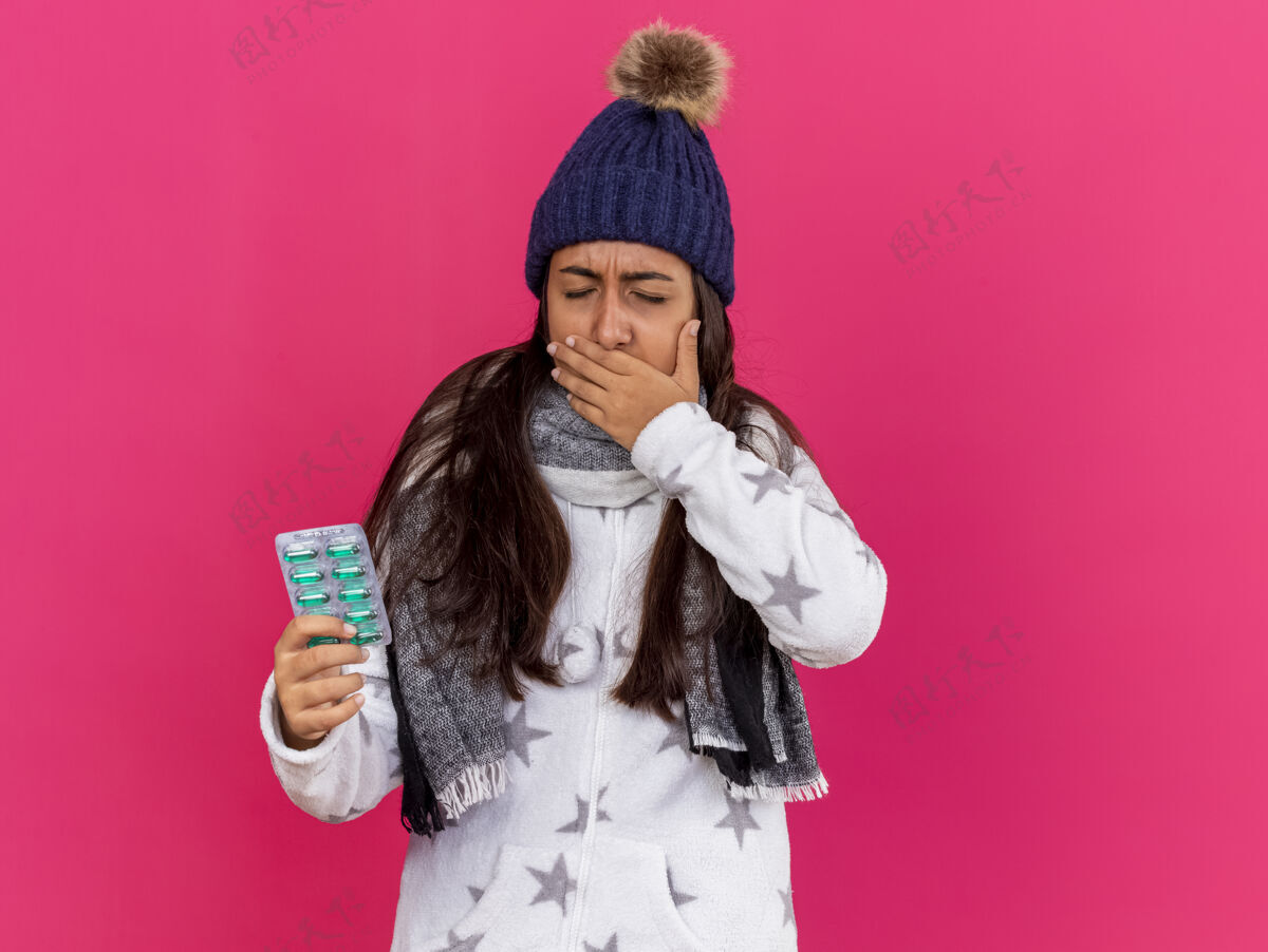 覆盖年轻的病女孩闭着眼睛戴着冬天的帽子戴着围巾拿着药丸手上包着酸痛的嘴隔离在粉色上疼痛持有嘴巴