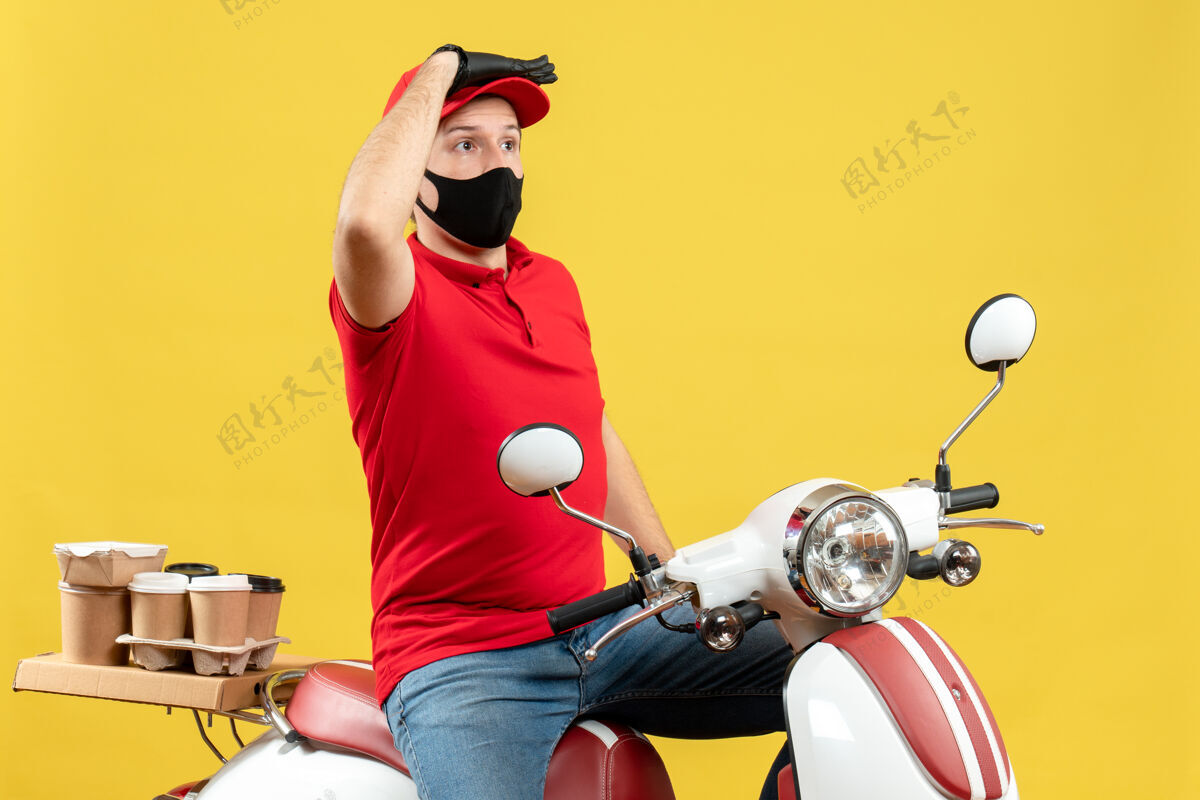 坐着戴着医用面罩 穿着红色上衣和帽子手套的年轻人坐在踏板车上传递命令的俯视图聚焦在黄色背景上的东西上交付东西命令