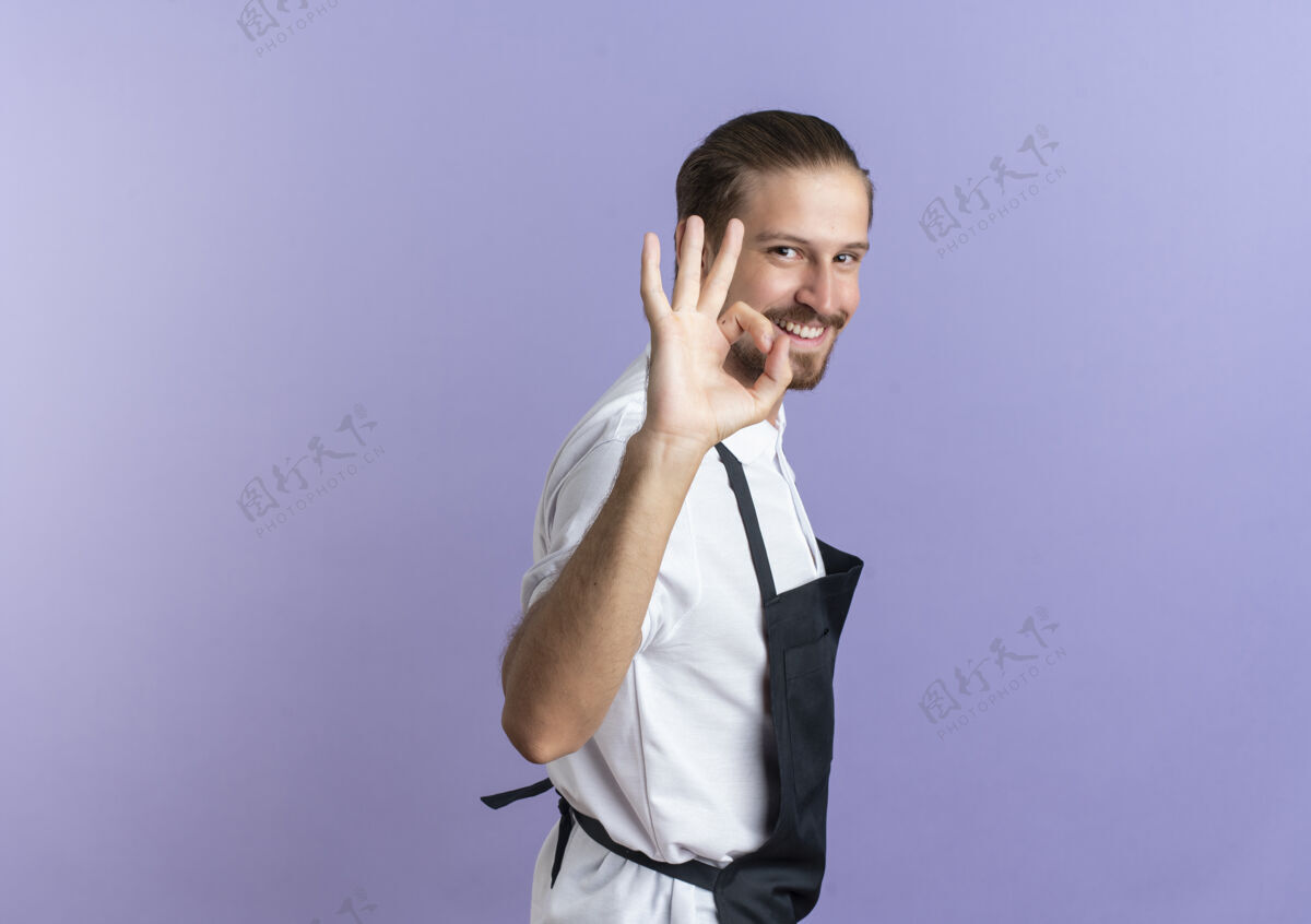脸面带微笑的年轻英俊的理发师穿着制服站在侧视图和做ok标志隔离在紫色的墙上站着感觉帅气