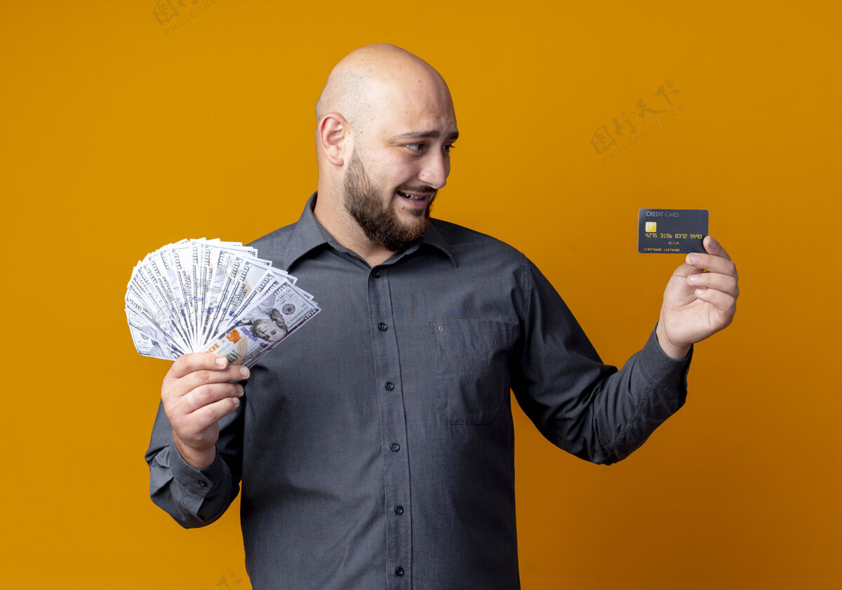 感情微笑着的年轻秃头呼叫中心男子拿着信用卡和钱 看着隔离在橙色墙上的卡中心橙色年轻